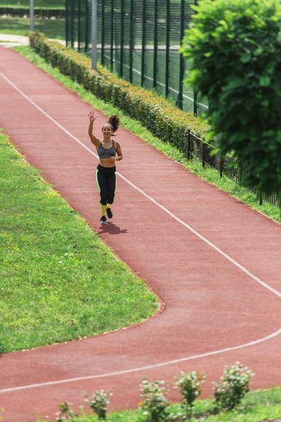 pełna długość szczęśliwy sportowiec w słuchawki bezprzewodowe machając ręką podczas biegania na stadionie - Zdjęcie, obraz
