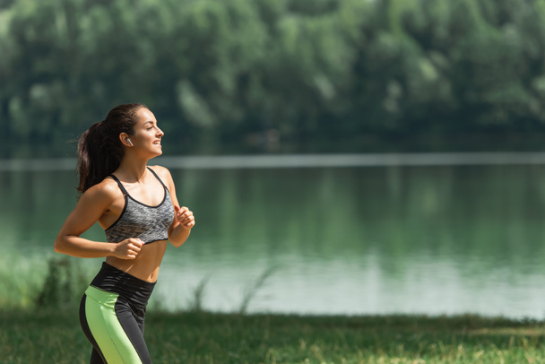 Seitenansicht einer glücklichen Sportlerin mit drahtlosen Kopfhörern, die Musik hört, während sie im grünen Park am See läuft - Foto, Bild