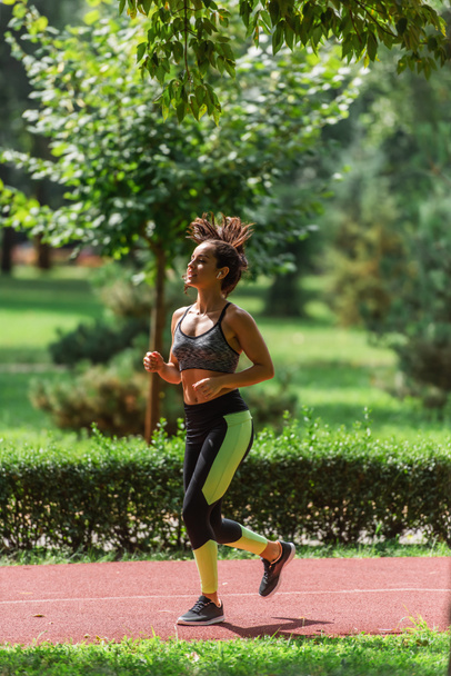 full length of young athletic woman in wireless earphones running on path in green park - 写真・画像