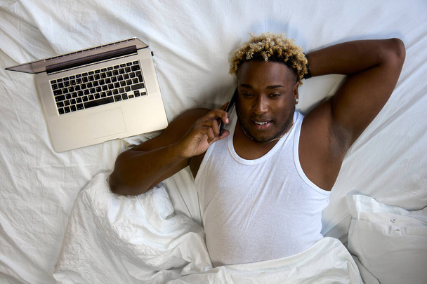 Top view of a black man lying on a snow-white bed next to a modern laptop and talking on a mobile phone - Foto, afbeelding