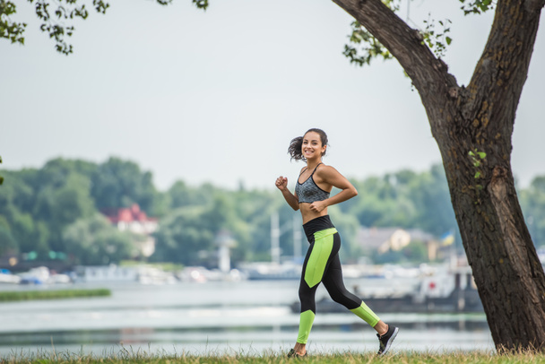 full length of joyful sportswoman in crop top and leggings jogging in park near lake - Photo, Image