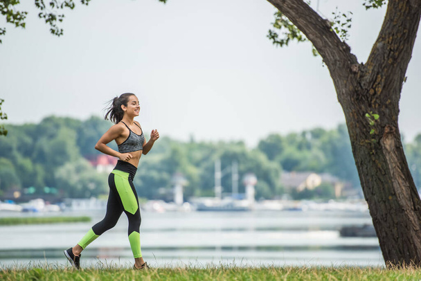 full length of cheerful sportswoman in crop top and leggings jogging in park near lake - Foto, Bild