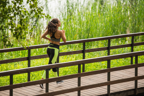 full length of happy sportswoman in crop top and leggings jogging on bridge in park  - Fotó, kép