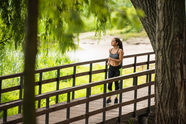 公園の湖の近くの橋の上をジョギングする作物のトップとレギンスの幸せなスポーツ女性の完全な長さ  - 写真・画像