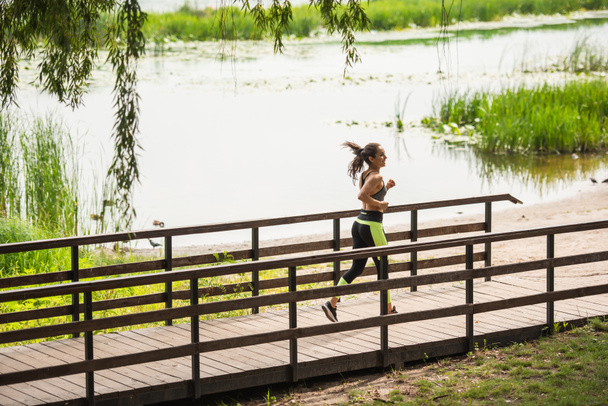 full length of happy woman in crop top and leggings jogging on bridge near lake in park  - Φωτογραφία, εικόνα
