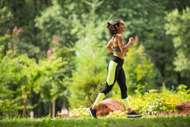 volledige lengte van vrolijke sportvrouw in gewas top en leggings joggen op gras in park  - Foto, afbeelding