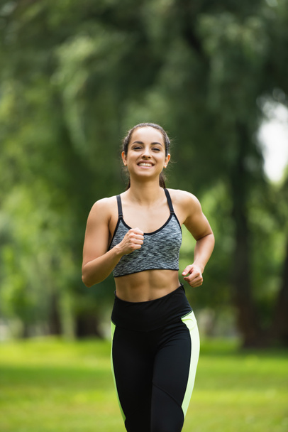 joyful young woman in crop top and leggings jogging in park  - Photo, Image