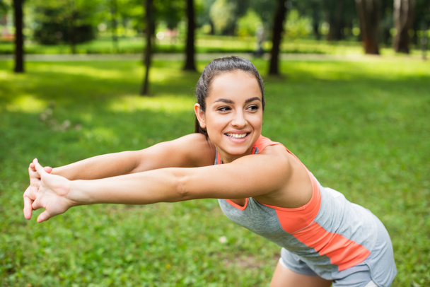 joyful and sportive woman stretching while warming up in park  - Zdjęcie, obraz