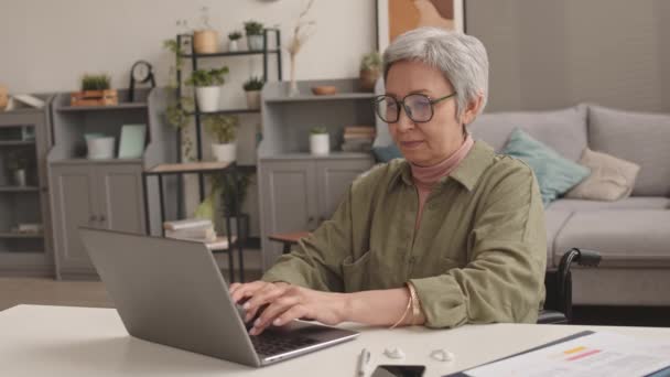 車椅子のアジア系のインテリジェントシニア女性の中,自宅の職場で机の上にポータブルコンピュータを入力します - 映像、動画