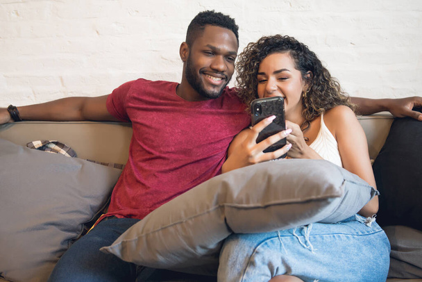Couple using a mobile phone while sitting on a couch at home. - Fotoğraf, Görsel
