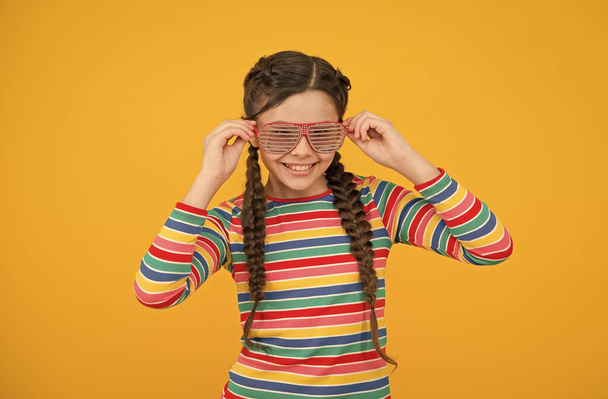 Saçları örgülü mutlu genç kız sarı arka planda gökkuşağı renginde ve göz kamaştırıcı parti gözlükleri takıyor. - Fotoğraf, Görsel