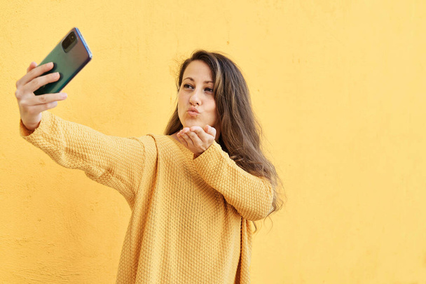 Porträt der schönen Mädchen schießen Selbstporträt auf Smartphone-Kamera bläst Luftkuss isoliert auf gelbem Hintergrund. Konzept der Technologie - Foto, Bild