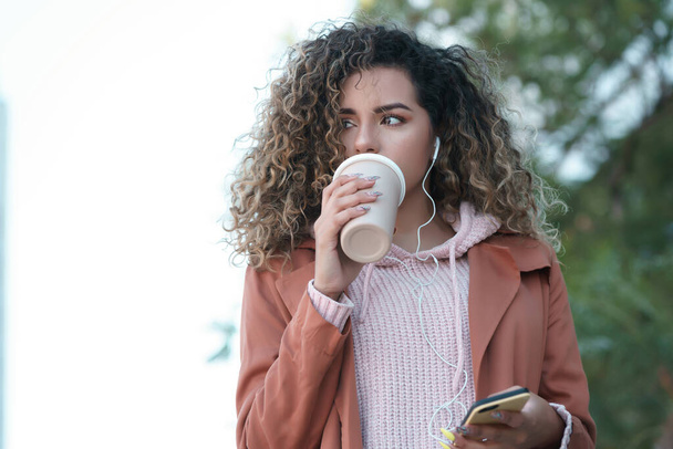 Femme latine boire du café et écouter de la musique avec son téléphone à l'extérieur. - Photo, image