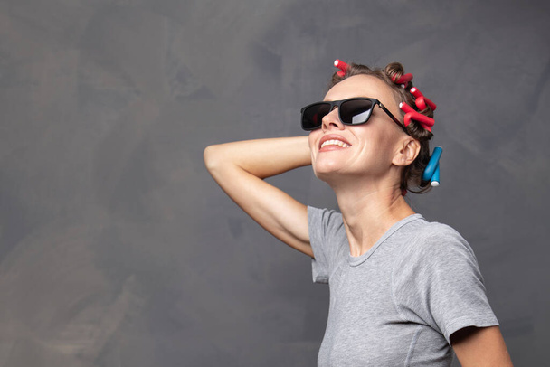 portrait of a young funny woman wearing sunglasses, t shirt and curlers over grey background, cop space - 写真・画像