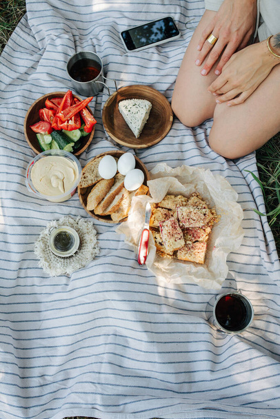 Picnic meal on a blanket. Top view. Girl sitting on her legs in a corner. No face. Tea, eggs, bread, hummus, vegetables, cake. - Zdjęcie, obraz