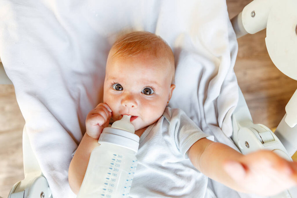 Мила маленька новонароджена дівчинка п'є молоко з пляшки і дивиться на камеру на білому тлі. Немовля, що їсть молоко, лежить на головному стільці вдома. Батьківство щаслива дитина
 - Фото, зображення