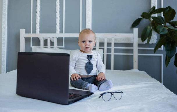 Takım elbiseli küçük bir çocuk dizüstü bilgisayarla yatakta oturuyor. - Fotoğraf, Görsel