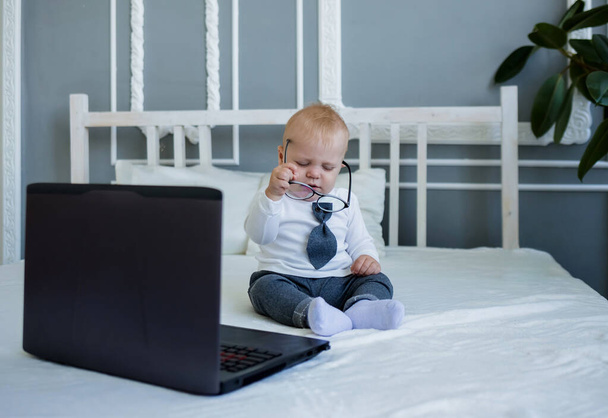 Takım elbiseli ve gözlüklü küçük çocuk dizüstü bilgisayarla yatakta oturuyor. - Fotoğraf, Görsel