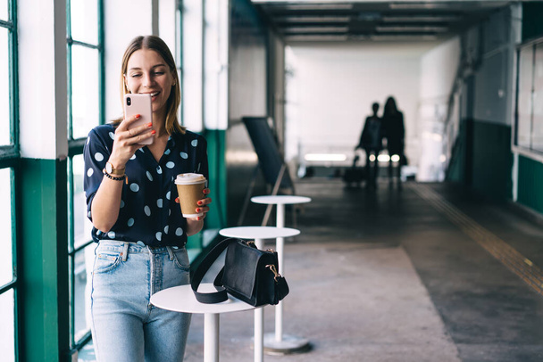 Happy caucasian blonde hipster girl reading message from social networks on mobile phone during coffee break, positive 20s woman in trendy wear watching videos and chatting via smartphone and 4G - Foto, Imagen