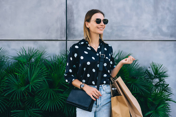 Positive beautiful 20s woman in fashion clothes holding shopping bags with items on sales in store, cheerful blonde caucasian female consumer in stylish spectacles enjoying buying and shopping - Foto, Bild