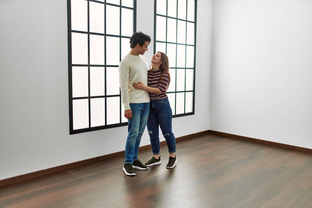 Jovem casal hispânico sorrindo feliz e abraçando de pé em casa nova vazia. - Foto, Imagem