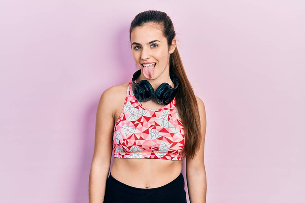 Jonge brunette tiener het dragen van sportschool kleding en het gebruik van koptelefoon steken tong uit blij met grappige uitdrukking. emotie concept.  - Foto, afbeelding