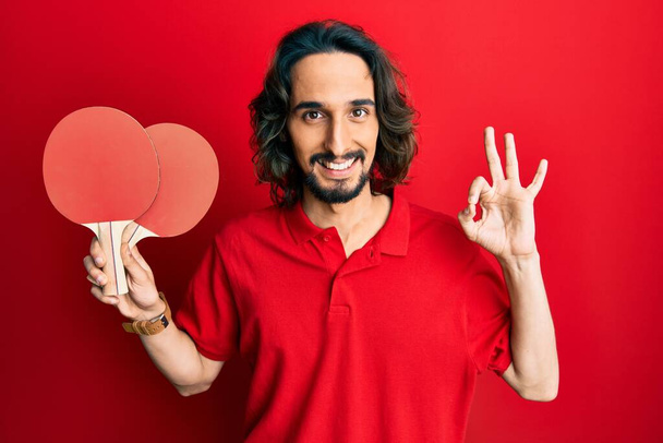 Fiatal spanyol férfi kezében piros ping-pong ütők csinál rendben jel ujjakkal, mosolygós barátságos gesztus kiváló szimbólum  - Fotó, kép