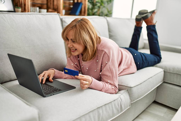 Femme blonde du moyen âge achetant à l'aide d'un ordinateur portable et d'une carte de crédit couchée sur le canapé à la maison. - Photo, image