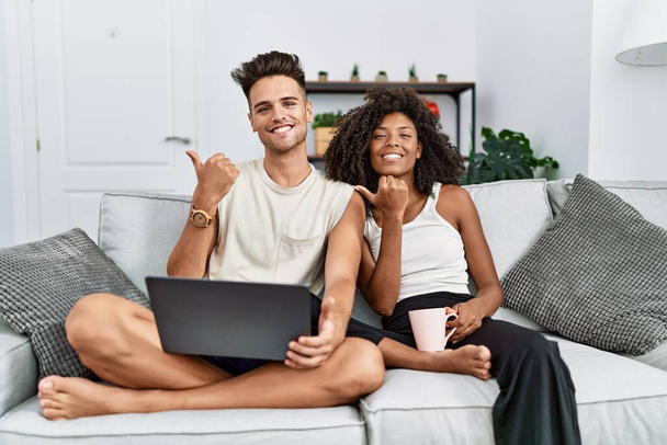 Junge interrassische Paar mit Laptop zu Hause sitzt auf dem Sofa und zeigt nach hinten mit der Hand und Daumen nach oben, lächelt zuversichtlich  - Foto, Bild