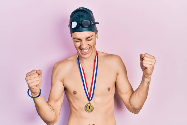 Young hispanic man wearing swimmer glasses and gold medal very happy and excited doing winner gesture with arms raised, smiling and screaming for success. celebration concept.  - Photo, image
