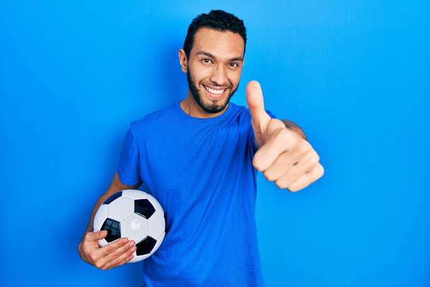 Hispanic man with beard holding soccer ball approving doing positive gesture with hand, thumbs up smiling and happy for success. winner gesture.  - Photo, Image