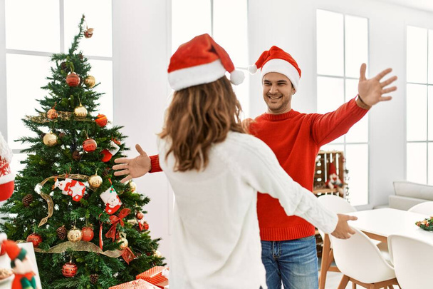 Joven pareja hispana con sombrero de navidad sonriendo feliz con los brazos abiertos para abrazarse en casa. - Foto, Imagen