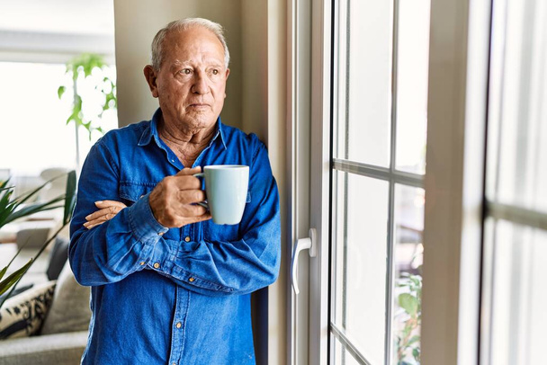 Ένας ηλικιωμένος άνδρας με γκρίζα μαλλιά να γέρνει στο παράθυρο του σπιτιού του, πίνοντας ένα φλιτζάνι καφέ το πρωί. - Φωτογραφία, εικόνα