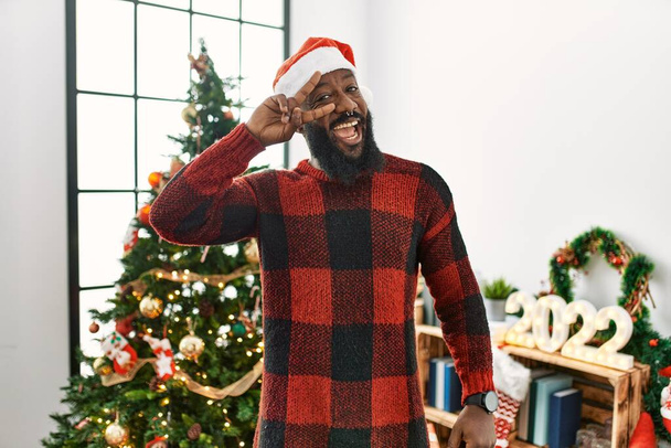 Αφροαμερικάνος που φοράει το καπέλο του Άη Βασίλη στέκεται δίπλα στο χριστουγεννιάτικο δέντρο κάνοντας το σύμβολο της ειρήνης με τα δάχτυλα πάνω από το πρόσωπο, χαμογελώντας χαρούμενα δείχνοντας νίκη  - Φωτογραφία, εικόνα