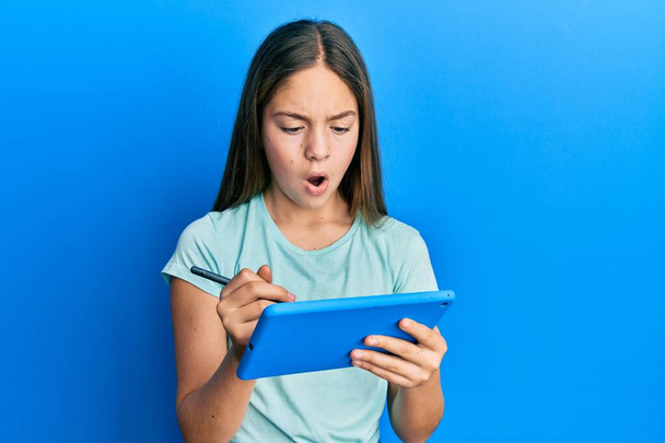 Beautiful brunette little girl using touchpad drawing on screen in shock face, looking skeptical and sarcastic, surprised with open mouth  - Fotoğraf, Görsel