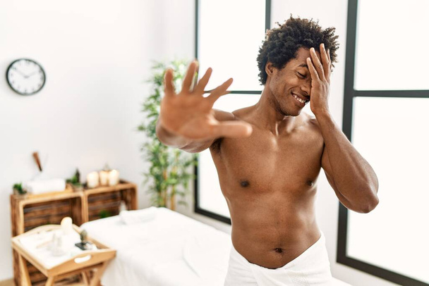 Jonge Afro-Amerikaanse man shirtloos dragen handdoek staan op schoonheidscentrum bedekken ogen met handen en doen stop gebaar met droevige en angst uitdrukking. beschaamd en negatief concept.  - Foto, afbeelding