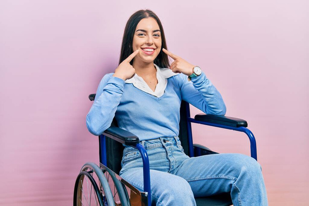 Hermosa mujer con ojos azules sentada en silla de ruedas sonriendo con la boca abierta, los dedos señalando y forzando la sonrisa alegre  - Foto, Imagen