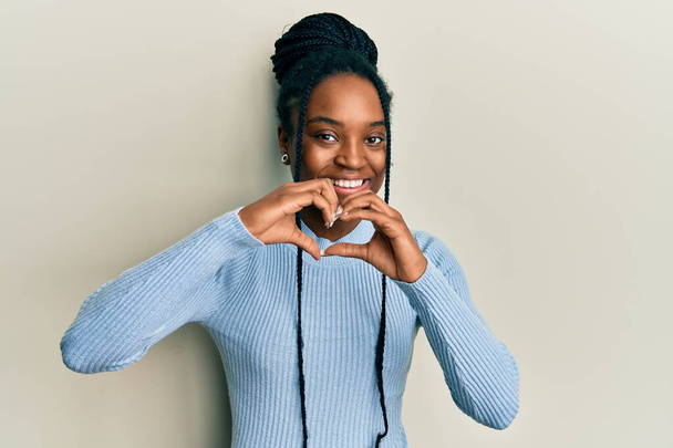 手で心のシンボル形状を行う愛に笑みを浮かべてカジュアルな青いセーターを着て編組髪を持つアフリカ系アメリカ人女性。ロマンチックなコンセプト.  - 写真・画像