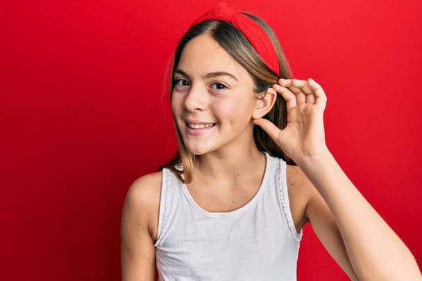 Beautiful brunette little girl wearing casual white t shirt smiling with hand over ear listening and hearing to rumor or gossip. deafness concept.  - Photo, Image