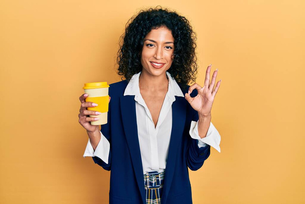 Jong latijn meisje draagt zakelijke stijl drinken kopje koffie doet ok teken met de vingers, glimlachend vriendelijk gebaar uitstekend symbool  - Foto, afbeelding