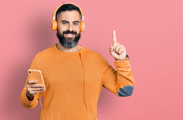 Latinomies, jolla on parta älypuhelimella yllään kuulokkeet yllättynyt ideasta tai kysymyksestä, joka osoittaa sormella onnellisia kasvoja, numero yksi  - Valokuva, kuva