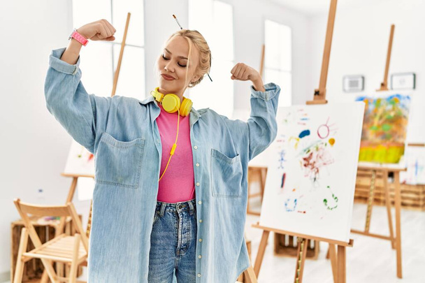 Joven chica caucásica en el estudio de arte mostrando los músculos de los brazos sonriendo orgulloso. concepto de fitness.  - Foto, imagen