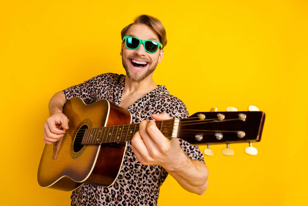 Фото милого милого молодого джентльмена в печатной рубашке темные очки, играющие гитарное пение изолированный желтый цвет фона - Фото, изображение