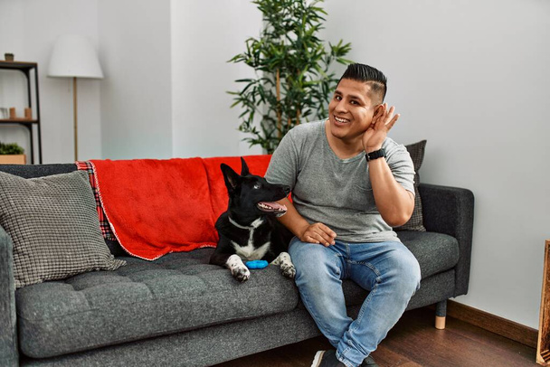 Fiatal latin férfi és kutya ül otthon a kanapén, és fültől fülig mosolyogva hallgatják a pletykát vagy pletykát. süketség fogalma.  - Fotó, kép