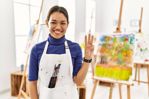 Mladá brunetka žena v ateliéru umění ukazuje a ukazuje prsty číslo tři a zároveň se usmívá sebevědomě a šťastně.  - Fotografie, Obrázek