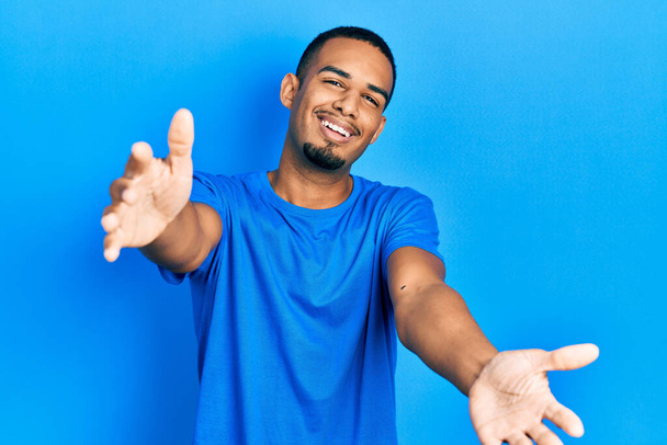 Ein junger afrikanisch-amerikanischer Mann trägt ein lässiges blaues T-Shirt und blickt lächelnd mit offenen Armen in die Kamera, um sich zu umarmen. fröhlicher Ausdruck, der das Glück umarmt.  - Foto, Bild