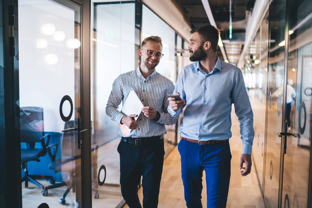 Positive male entrepreneurs in smart casual wear strolling in office using technology talking to each other, smiling prosperous men business partners satisfied with cooperation and meeting in office - Foto, Imagem