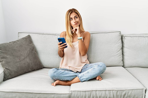 Blonde schöne junge Frau, die zu Hause auf dem Sofa sitzt und ihr Smartphone benutzt, mit der Hand am Kinn, über Fragen nachdenkend, nachdenklich. Lächeln mit nachdenklichem Gesicht. Zweifelhaftes Konzept.  - Foto, Bild