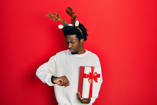 Giovane uomo afroamericano che indossa il cappello di Natale dei cervi che tiene il regalo controllando il tempo sull'orologio da polso, rilassato e fiducioso  - Foto, immagini