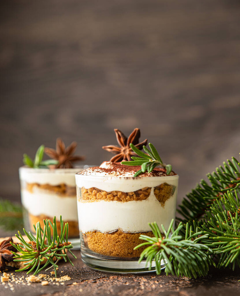 Layered dessert in glass jar with cookie crumble and whipped cream decorated with rosemary and anise, dark brown background. No bake cheesecake, trifle or pudding. - Fotoğraf, Görsel